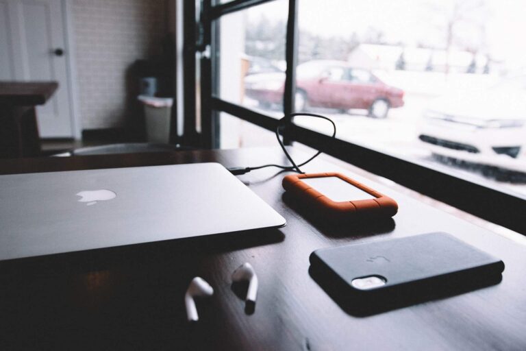 Closed silver MacBook on a dark desk with an orange rugged external drive and an iPhone nearby by a window edge.