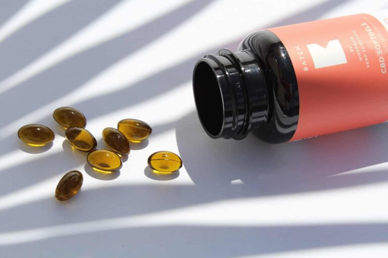 Orange CBD oil bottle tipped over with scattered amber capsules on a white surface under striped light.