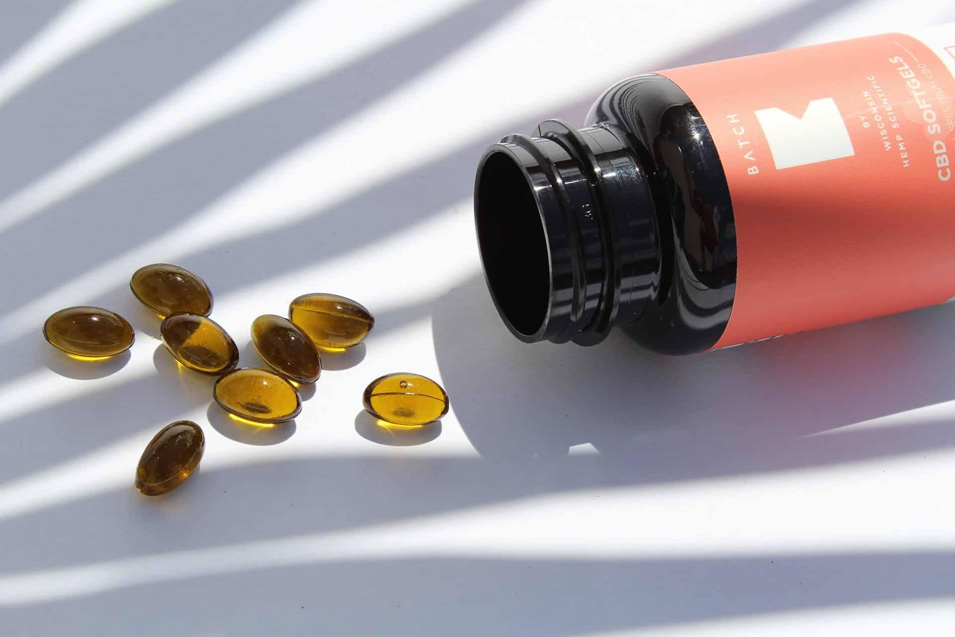 Orange CBD oil bottle tipped over with scattered amber capsules on a white surface under striped light.