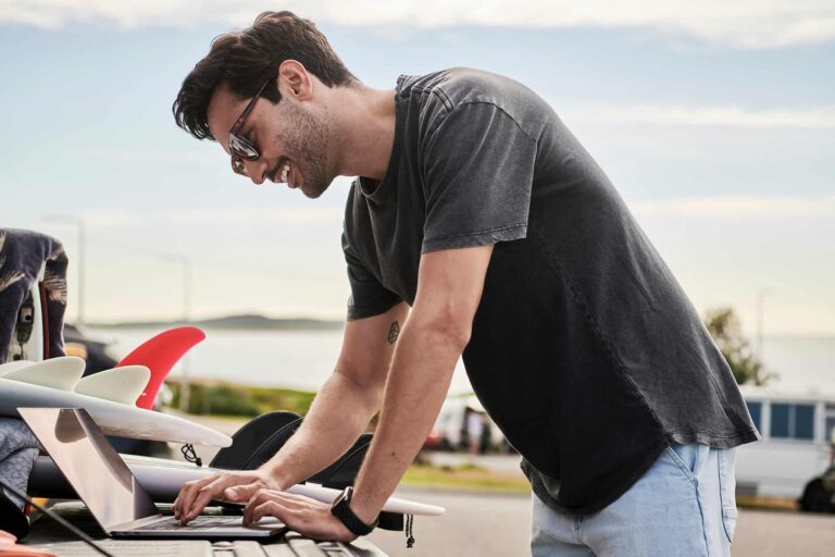 Man wearing sunglasses and a dark T-shirt leans over a laptop on a surfboard rack outdoors, near the beach on a sunny day.