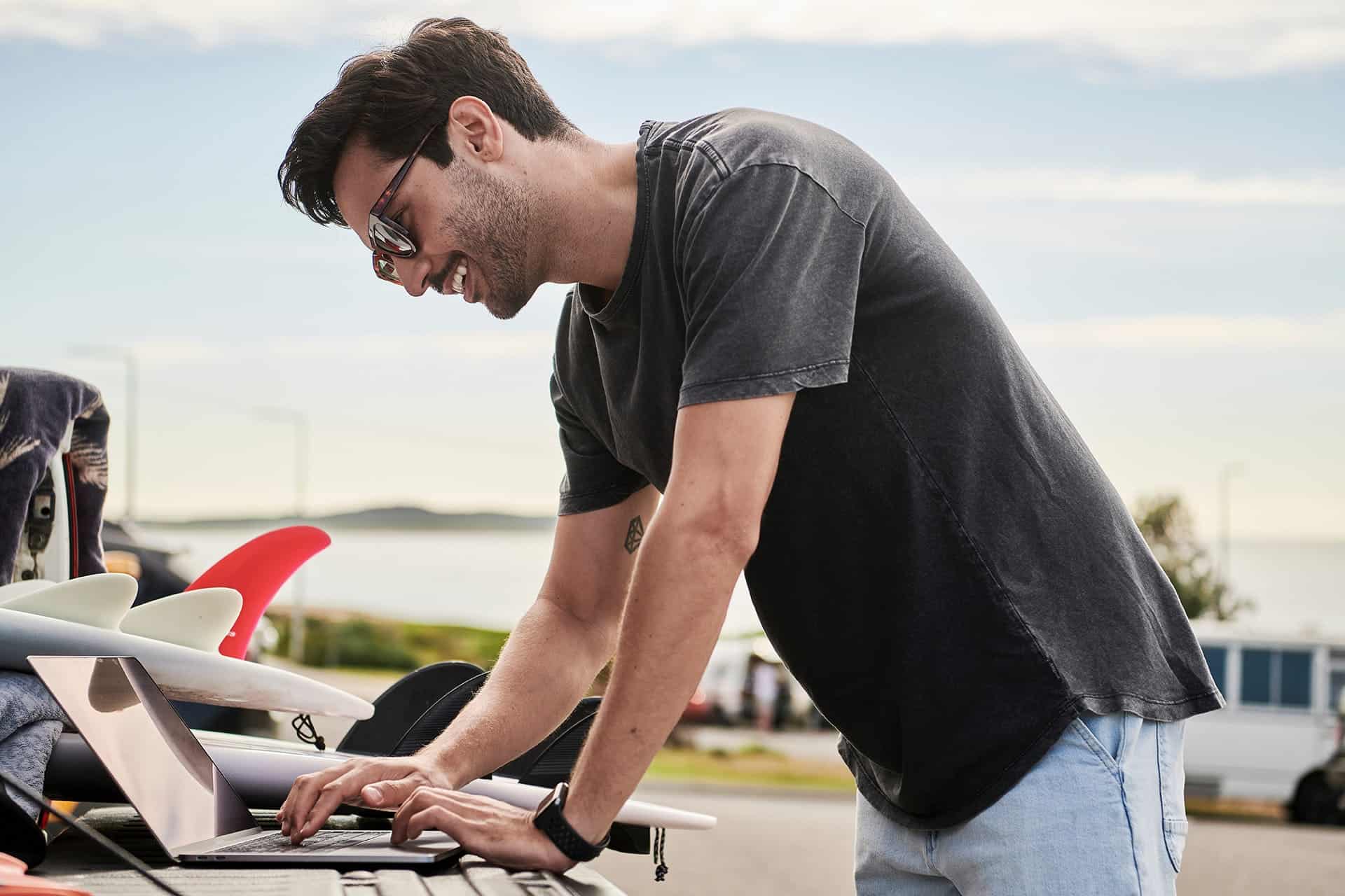 Man wearing sunglasses and a dark T-shirt leans over a laptop on a surfboard rack outdoors, near the beach on a sunny day.