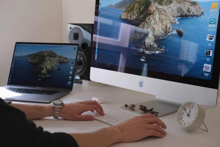 Person typing on a white keyboard at a desk with a large iMac displaying an ocean wallpaper; a laptop, watch, sunglasses, and a clock nearby.