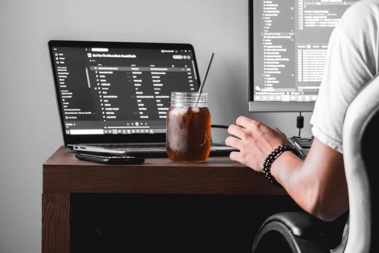 Person seated at a wooden desk with a laptop displaying code/music app, a glass mason jar iced drink with a straw in front, and a large monitor showing more code in the background.