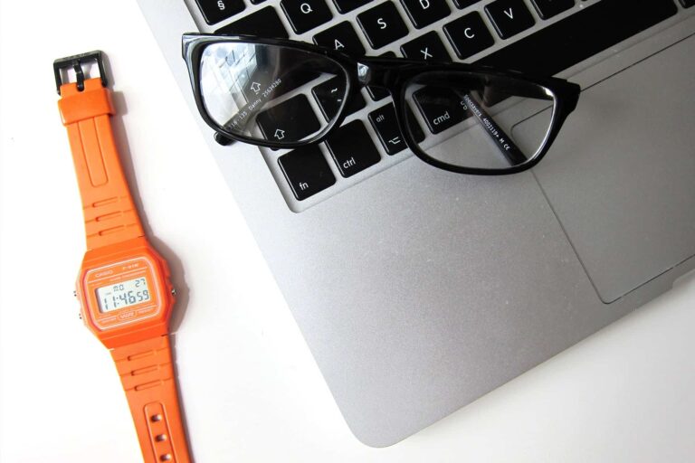 Orange Casio digital wristwatch placed beside a laptop keyboard with black eyeglasses resting on the keys.
