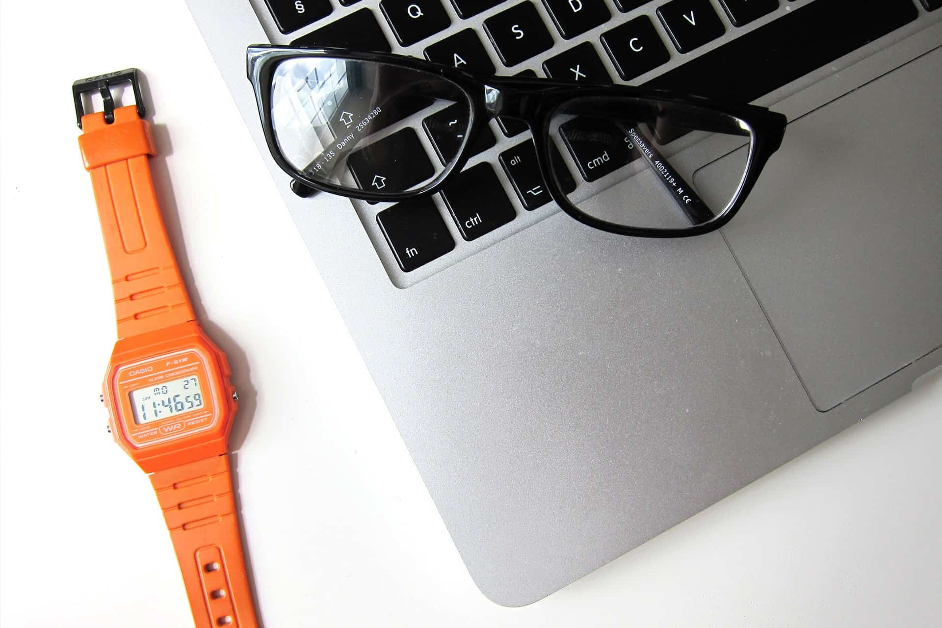 Orange Casio digital wristwatch placed beside a laptop keyboard with black eyeglasses resting on the keys.