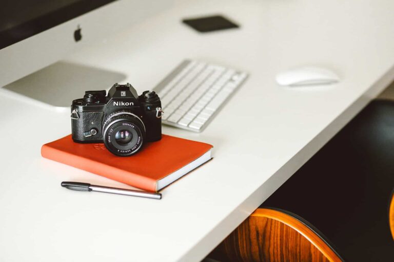 Nikon camera sits on an orange notebook on a white desk, with a keyboard and mouse blurred in the background.