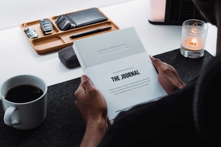Person holding open magazine titled The Journal at a desk, with a coffee mug nearby and a lit candle in the background.