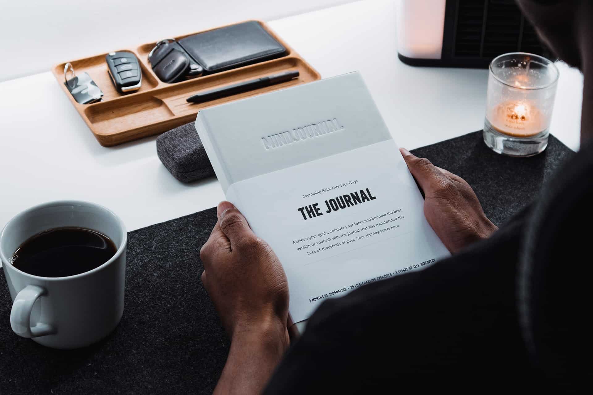 Person holding open magazine titled The Journal at a desk, with a coffee mug nearby and a lit candle in the background.