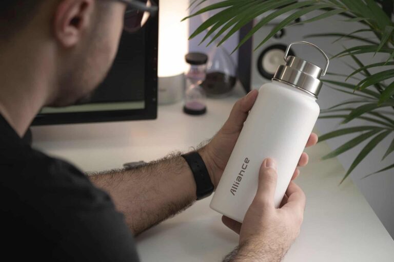 Person holding a white reusable water bottle with a metal cap at a desk with a plant nearby.