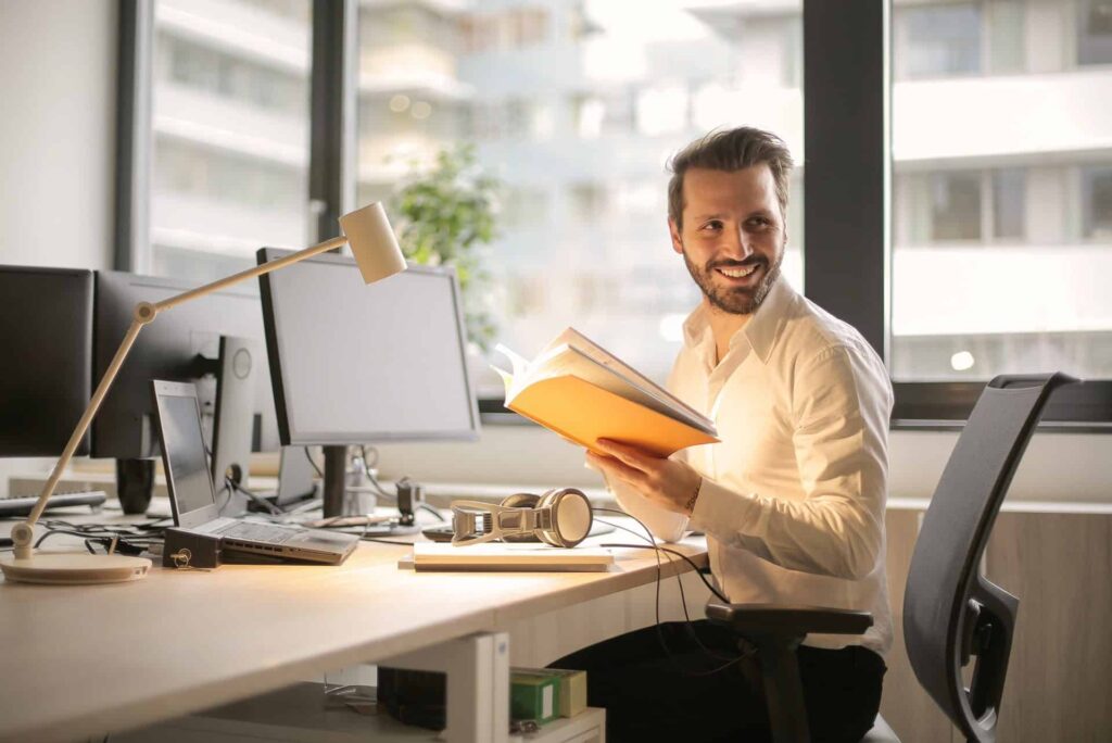 Man in a white shirt sits at a bright office desk, smiling while holding an orange notebook.