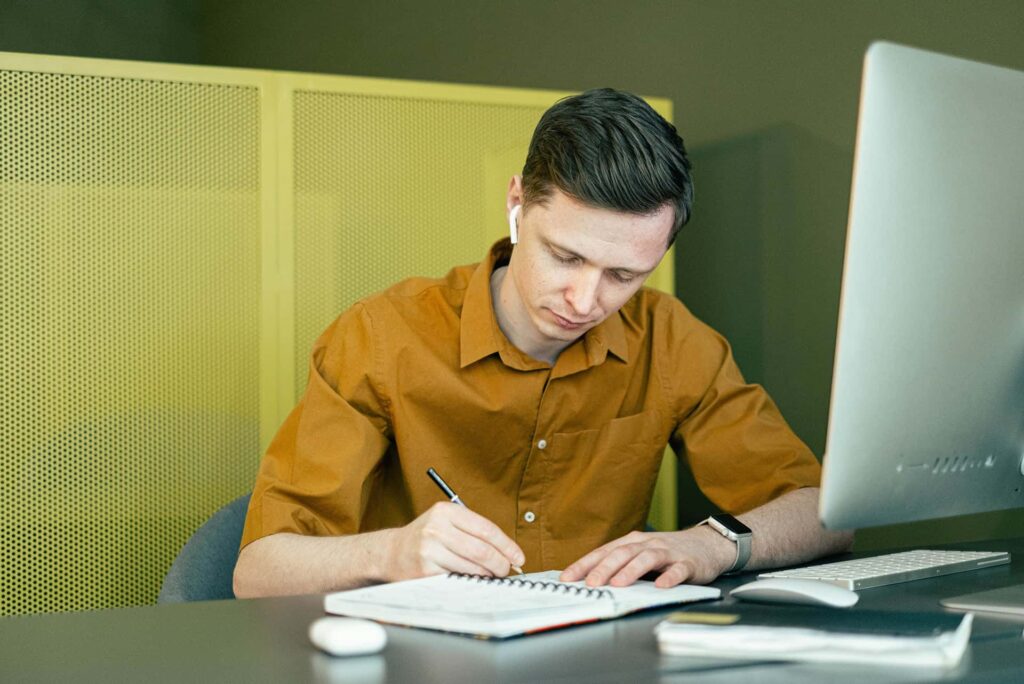 Man wearing a brown shirt writes in a spiral notebook at a desk, with a computer monitor nearby and wireless earbuds in his ear.