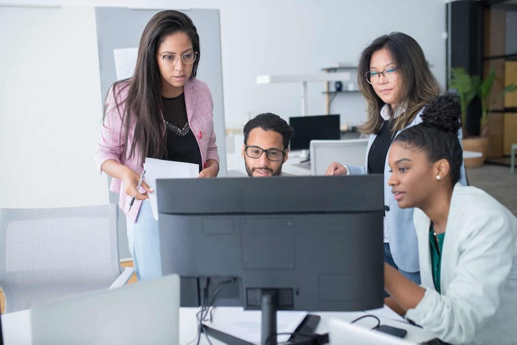 Group of four coworkers collaborating around a desk, looking at a computer monitor in a bright office.