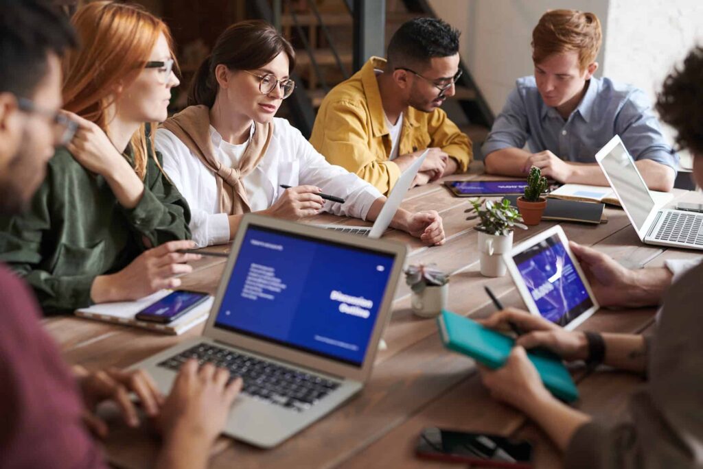 Group of professionals collaborating around a wooden table with laptops and tablets on a shared workspace.