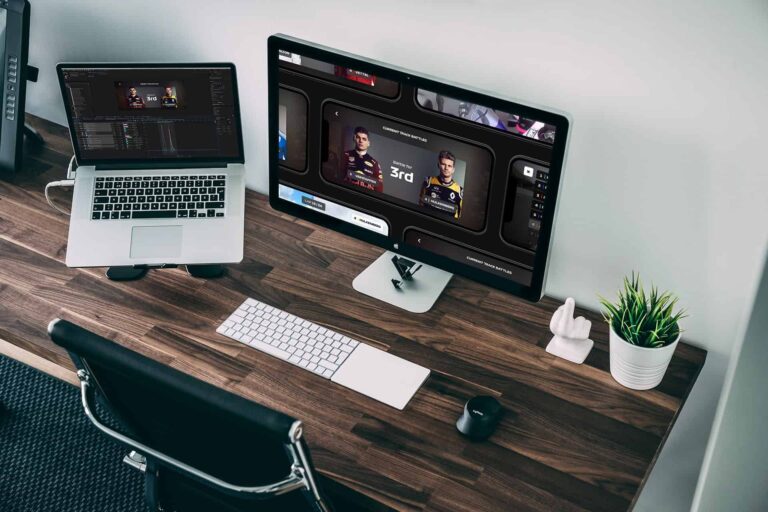 Modern workspace with a laptop and a large monitor showing a game/dashboard on a wooden desk.