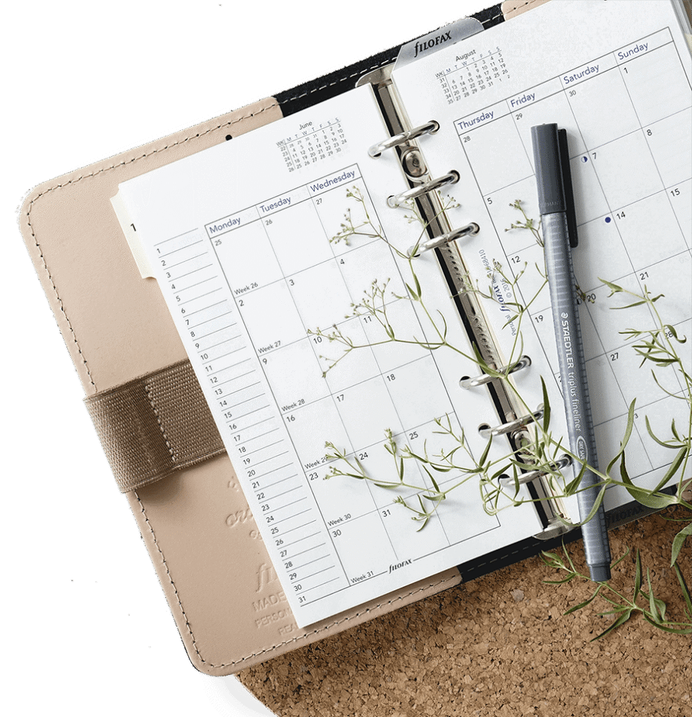 Open planner with monthly calendar pages, a gray marker, and sprigs resting on a cork board surface.
