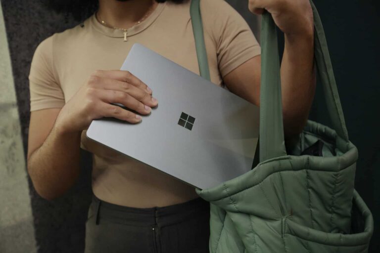 Person in a beige top holds a silver-gray laptop with a four-square Windows logo against a green backpack.