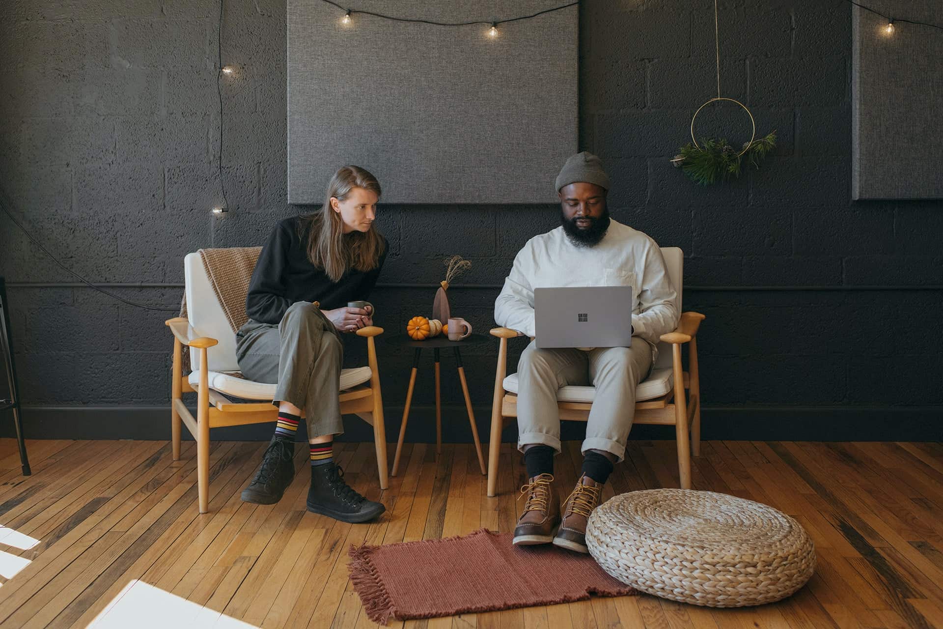 Two people sit on wooden chairs in a cozy dark-walled room; man uses a laptop while woman holds a mug with autumn pumpkins on the table nearby.