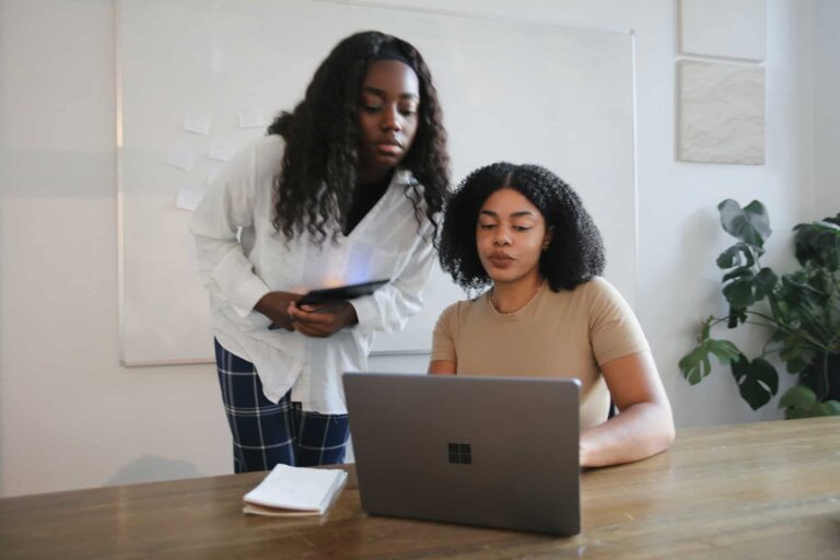 Two women collaborate in an office: one standing with a tablet, the other seated at a laptop near a whiteboard and plant.