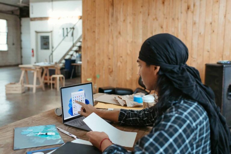 Person wearing a black head wrap uses a laptop at a wooden table in a bright workshop.