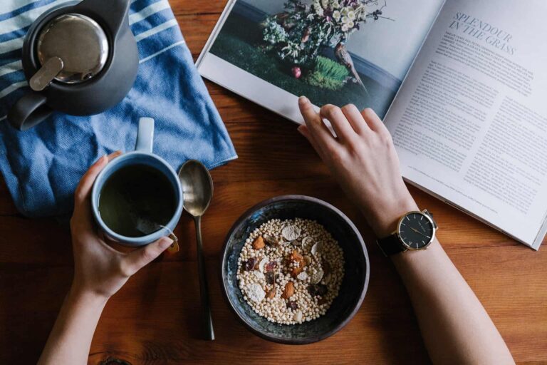 Top-down view of a breakfast setup: hands hold a blue mug over a bowl of granola on a wooden table with an open magazine nearby.