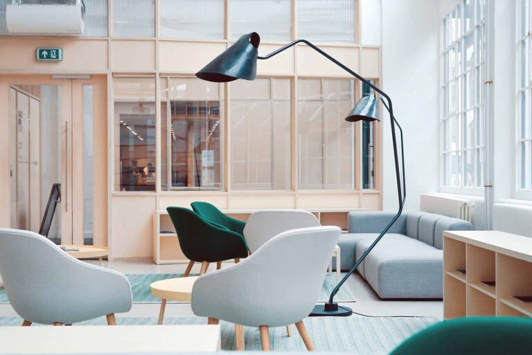 Bright modern office lounge with pale blue chairs, a gray sofa, and a large black arc floor lamp near a sunlit windowed wall.